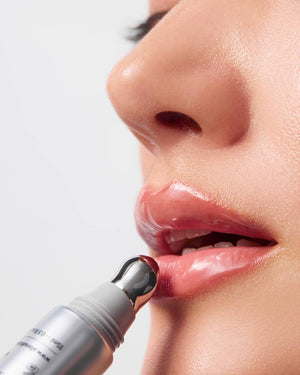 Close-up of a person's lower face applying a silver cosmetic tube with a reflective gel tip to the lips against a light gray background.
